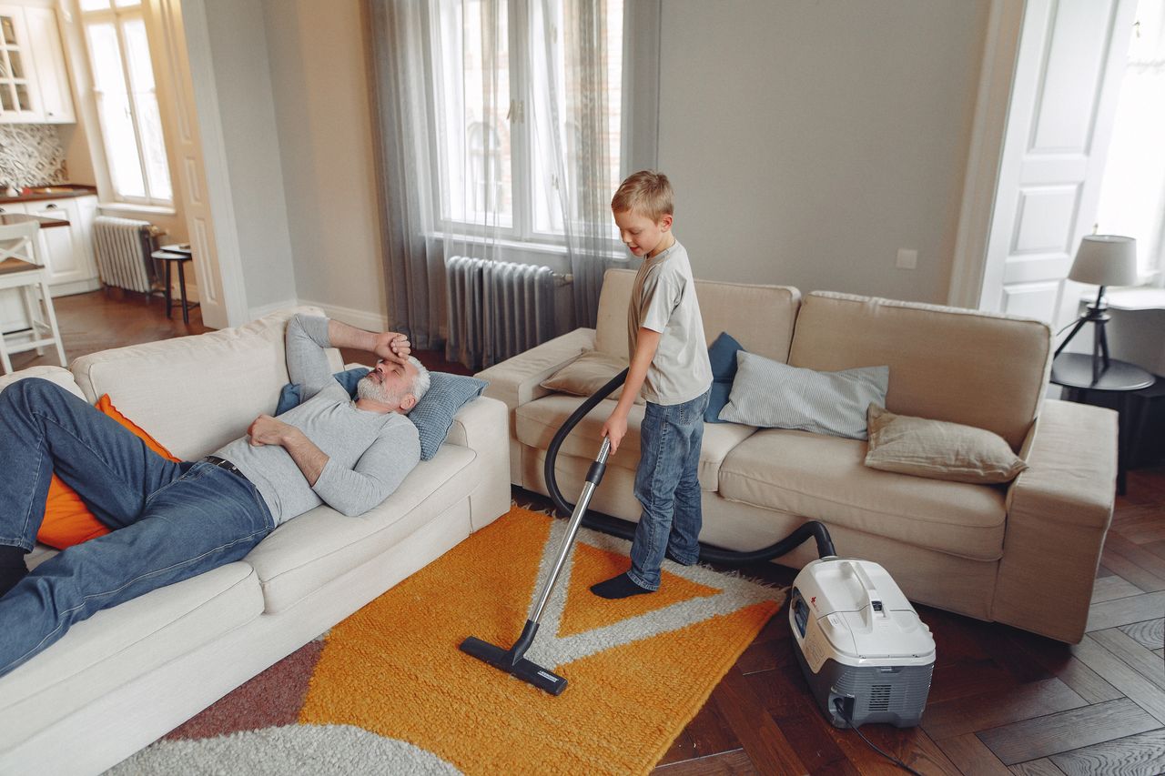 A young boy is vacuuming the living room carpet while his grandfather rests on the sofa.
